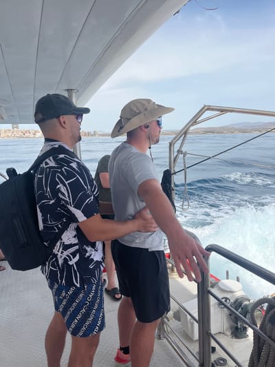 Two men are standing on the deck of a boat, looking out at the ocean. The man on the left is wearing a black and white patterned shirt, blue and white patterned shorts, a dark baseball cap, and sunglasses. He has a black backpack on his back. The man on the right is wearing a light grey t-shirt, black shorts, a tan bucket hat, and sunglasses. He has his left arm around the waist of the man on the left. The boat is moving, as evidenced by the wake behind it. In the distance, a city skyline is visible along the coast. The sky is blue with some clouds. The scene appears to be taking place in the Mediterranean Sea, possibly near Elx/Elche, Spain, given the architecture of the distant city.