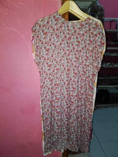 A floral patterned dress, predominantly gray with red and pink floral accents, hangs on a wooden hanger against a pink wall. The dress appears to be made of a lightweight fabric. To the right of the dress, shelves are visible, containing what appear to be various items stored in plastic bags and some dark fabric. The floor in front of the shelves is tiled in a blue-gray color. The environment suggests an indoor setting, possibly a shop or storage area, located in Sendangrejo, Indonesia. There are no people visible in the image.