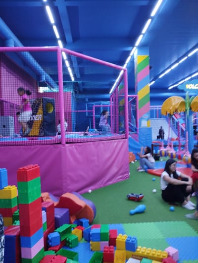 The image depicts an indoor children's play area in Talas City, Kyrgyzstan. The environment is brightly lit with fluorescent lights from the blue ceiling. The floor is covered in green artificial turf, with blue and yellow foam mats arranged in sections. Prominently featured are large, colorful building blocks, resembling oversized LEGO bricks, in red, blue, green, and yellow.
A pink, netted play structure dominates the left side of the frame, with children visible inside. One child in a pink shirt and white shorts is in motion within the structure. Various soft play elements, including orange and purple foam shapes, are scattered on the floor.
Several individuals, primarily women and girls, are present in the play area. Some are seated on the floor, observing or resting. One woman in a white shirt and black pants is looking to her right, appearing to engage in conversation. Another woman, partially visible in the lower right, is wearing glasses.
The background reveals more of the play facility, including colorful slides and hanging spherical play elements. A sign with the word "VOLCANO" is partially visible on a yellow structure in the upper right. A red toy car and a blue plastic object are on the green floor. The overall impression is of a vibrant and active recreational space for children.