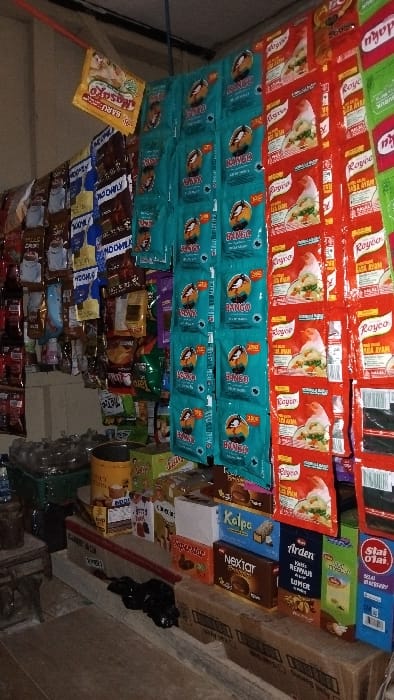 The image shows a small shop located in the city of Kecamatan Panjalu, Indonesia, with coordinates -7.143 latitude and 108.278 longitude. The shop is stocked with various food items, including packaged snacks, instant coffee, and chocolate bars. The shelves are organized with different brands and products. Some of the products visible on the shelves are "Royco" for soup and "Bango" for soy sauce. The shop also has a few boxes of "Nectar" chocolate bars and a box of "Slai O'lai" crackers. The shop is lit by natural light coming from the open door. There are no people in the scene, and the shop appears to be empty. The floor is made of wooden planks.  The shelves are in a wooden frame and appear to be slightly damaged.  The shop is located in a residential area with a brick wall behind the counter.