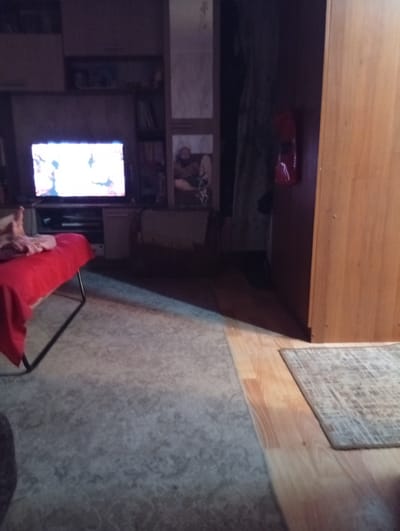 The image depicts an indoor scene, possibly a living room or bedroom, with low lighting. A television screen is illuminated with a bright, abstract image. To the left, a person's legs and feet are visible, resting on a red blanket draped over what appears to be a chaise lounge or a low bed. The floor is covered with two different rugs; one is a light gray with a speckled pattern, and the other is a lighter, wood-like laminate with a smaller, patterned rug placed on top of it. In the background, there are shelves with various items and a wooden wardrobe. The overall impression is of a dimly lit, lived-in space. No specific details or landmarks are visible in the image that would allow for a definitive identification of the location within Ruzaevka, Russia.