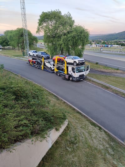 A car carrier truck is parked on the shoulder of a highway in Marchaux-Chaudefontaine, France. The truck is loaded with six cars, three on the top deck and three on the bottom deck. The cars are of various colors including white, gray, red, and black. The truck is white with a yellow hydraulic lift. The truck has a license plate that is partially visible. The highway is a multi-lane road with a concrete barrier separating the lanes. There is a grassy embankment on the left side of the highway, and trees and hills in the background. The sky is a pale pink and orange, suggesting it is either sunrise or sunset.