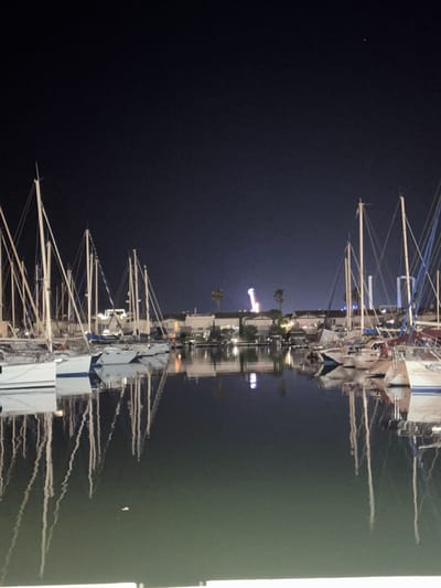 This image depicts a nighttime scene of a marina in the city of Agde, France. The foreground is dominated by the dark water of the marina, which reflects the lights from the boats and the distant cityscape. A number of sailboats are moored in the marina, their white hulls and masts clearly visible against the dark sky. The reflections of the boats are distorted by the ripples on the water, creating an abstract effect.
In the background, the lights of the city are visible, including the illuminated structures of what appears to be an amusement park or fairground. The sky is dark, with a few faint stars visible. The overall impression is one of a peaceful and serene night at the marina, with the city lights adding a touch of vibrancy to the scene.