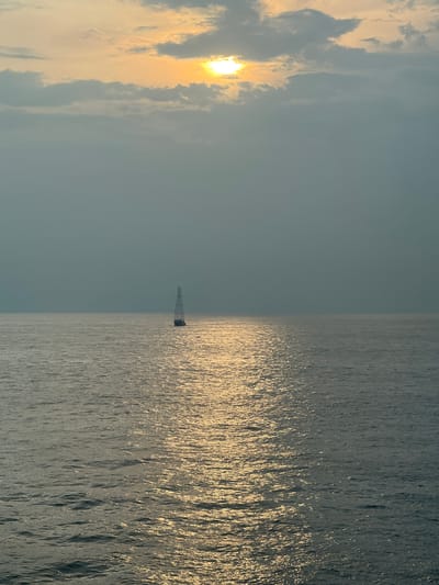 An aerial view captures a serene ocean scene at sunset. The sun, partially obscured by clouds, casts a golden shimmer across the water's surface, creating a reflective pathway. In the distance, a solitary sailboat with its mast raised gracefully glides across the calm sea. The horizon line is clearly defined, separating the vast expanse of water from a muted sky. The overall atmosphere is tranquil and picturesque, evoking a sense of peace and solitude. The image is devoid of any urban or specific geographical features, making the location indeterminate.