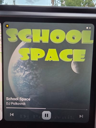 A close-up shot of a smartphone screen displays the album art for the song "School Space" by DJ Polkovnik. The artwork features the words "SCHOOL" stacked vertically next to "SPACE," both in a bright yellow, 3D block letter style. Behind the text is a cloudy image of a planet or moon. Playback controls are visible on the right, including a "pause" button. The location is most likely inside the apartment, as the background visible through the window implies that it's a sunny day outside. The city of Зеленая Роща-1 Киз, Russia is irrelevant.