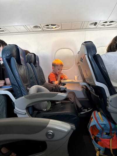 The image depicts the interior of an airplane cabin, with a young boy seated and looking at a tray table. The boy, with blonde hair and wearing an orange t-shirt, is seated in an aisle seat. In front of him, a small tray table is extended, holding a cup and some papers or a book. To his left, other passengers are partially visible, and to his right, another airplane seat is occupied by a person wearing a white shirt. A blue and orange backpack rests on the floor near the right side of the frame. The overhead compartments and air vents are visible at the top of the image. The scene is consistent with a typical airplane environment during a flight. The provided location "Vykhodnoi, Russia" is not discernible from the visual information within the image.