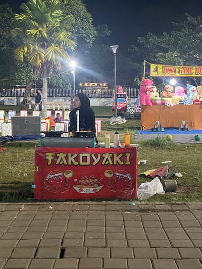 The image depicts an outdoor night scene, likely at a fair or market, in Sodong, Indonesia. In the foreground, a red banner displays the text "TAKOKYAKI" in yellow lettering, accompanied by cartoonish illustrations of octopuses. A person wearing a black hijab and black clothing stands behind a cooking station, seemingly preparing food. To the left of the food stand, various condiments in squeeze bottles are visible.
In the background, a tall palm tree with green fronds dominates the left side of the frame. Further back, a "ZONA GAME BO" sign is illuminated, suggesting a game area. Several large, colorful stuffed animals, including a pink bear and a blue character resembling Doraemon, are displayed on a counter, likely prizes for games. Other elements in the background include a street light, a metal fence, and trees. The ground in the foreground is paved with square grey tiles, while the middle ground and background consist of grassy areas. The overall ambiance is that of a lively evening event.