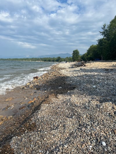 This image shows a rocky beach along a body of water, likely Lake Baikal, near Tolbazikha, Russia. The foreground is dominated by smooth, rounded pebbles in various shades of gray, brown, and white. Some of these rocks are wet from the lapping waves. The water, with its white foam and choppy surface, meets the shore at a gentle slope. Further inland, the beach transitions to sand, with a large piece of driftwood lying on it. In the background, a line of green trees forms a dense forest, beyond which mountains are visible under a cloudy sky. A few tents and what appear to be boats are sparsely scattered along the sandy shoreline, suggesting a recreational area. The overall atmosphere is tranquil and natural.