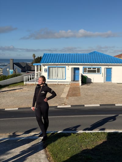 A person wearing a black wetsuit stands smiling warmly on a paved area next to a road, posing for the camera. They have one hand placed on their hip. The wetsuit features white branding, including the word "Mares" on the chest.
The scene is set in what appears to be a coastal town, likely within the Overstrand Local Municipality in South Africa. Behind the person is a two-story white building with a distinctive bright blue corrugated metal roof and blue window frames. A green refuse bin is visible near the building. In the background, the ocean stretches to the horizon under a clear blue sky with some scattered clouds. A palm tree is also visible, reinforcing the coastal setting.
The foreground shows a paved road with white and black curb markings, alongside a strip of green grass. Long shadows are cast across the road and grass, indicating either early morning or late afternoon sunlight. Visible text on a white sign attached to the building reads "SECURI-GUARD" with a partial phone number beginning "028 31...". The number "8" is also seen on the building. Another blue sign in the distance contains the text "THIS WAY TO..." and other unreadable details.