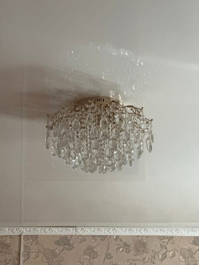 A close-up, low-angle shot shows a brass chandelier with cascading crystal droplets suspended from a white ceiling. The chandelier casts a dappled light pattern on the ceiling. Below the ceiling, a decorative border of molded white ornamentation separates the ceiling from patterned wallpaper. The wallpaper has a floral design in muted tones of beige and brown. The scene appears to be indoors, likely in a residential setting in Yazykova Pyatina, Russia, though no specific location details are visible within the frame itself.