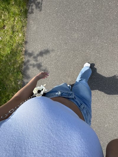 The image shows a first-person perspective of a person walking on an asphalt surface, potentially a sidewalk or path. The person is wearing blue jeans, white sneakers, and a light blue tank top with a silver chain. A shadow of the person is visible on the asphalt. The edge of a grassy area is seen on the left side of the image. The location is in Wassenberg, Germany.