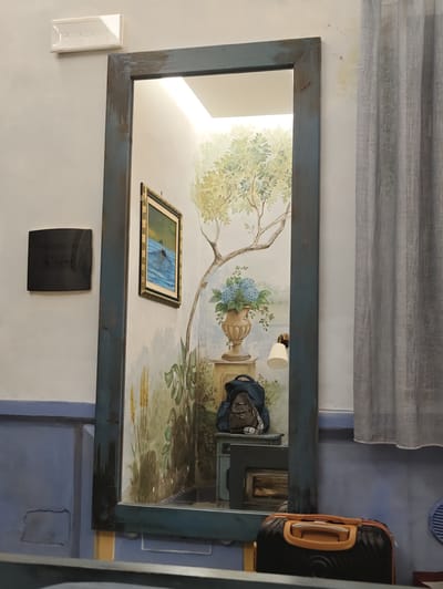 A full shot shows a large, distressed blue-framed mirror, reflecting a mural of a tree with blue flowers and a painting of a duck in water. To the left of the mirror, a black square object is mounted on the wall. Above it, a white rectangular light fixture is attached to the ceiling. A gray curtain hangs to the right of the mirror. In the foreground, a black and orange suitcase is partially visible, with a gray backpack placed on a blue table inside the mirror's reflection. The setting appears to be indoors, possibly a room or hallway in Pozzuoli, Italy, with blue and beige walls. No people are visible, and the lighting suggests it could be daytime or well-lit indoors.