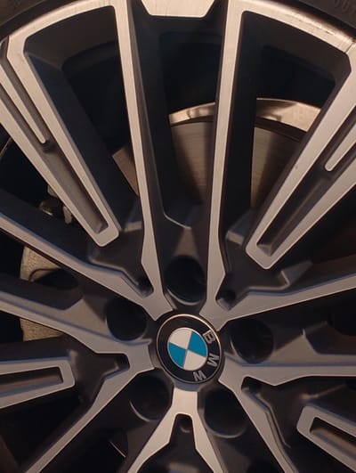 A close-up shot captures a BMW wheel with its characteristic logo. The wheel is made of dark grey alloy with silver accents and has a complex spoke design. The BMW logo, featuring the iconic blue and white circular emblem, is visible in the center of the wheel. Behind the wheel, a brake disc is partially visible. The overall image is focused on the wheel, highlighting its details and brand identity. The image does not provide any information about the city of Nor Geghi, Armenia.