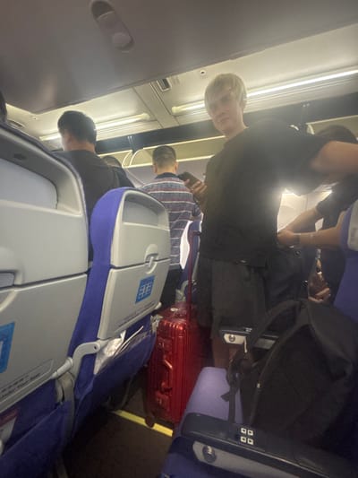 The image depicts the interior of an airplane cabin, likely in flight. The scene is crowded, with several passengers visible in the aisle and seated. In the foreground, a young man with blond hair, wearing a black t-shirt and dark shorts, is looking towards the camera and holding a smartphone. To his left, a red suitcase is placed in the aisle, blocking passage. Other passengers are seen further back, some appearing to be standing or moving within the confined space. The seats are upholstered in blue and gray fabric. The overhead compartments and lighting fixtures of the airplane are visible at the top of the frame. The specific location in Xinwu District, China, cannot be determined from the image alone.