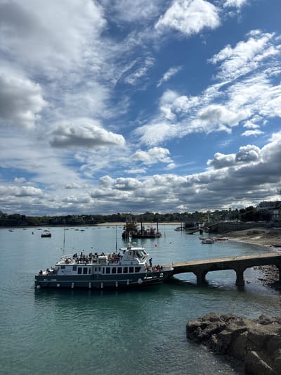 The image shows a large ferry boat docked at a concrete pier in Dinard, France. The boat is named "Corsaire II" and has many passengers onboard. It appears to be leaving the dock as people are walking up the gangplank to disembark. The pier is connected to a small rocky island by a pedestrian walkway. The water is turquoise and there are several other boats in the distance.  The sky is blue with a few white clouds. In the background, there are trees and buildings along the shoreline.  The location is at the port of Dinard, near the mouth of the Rance River, where the ferry departs for the nearby island of Saint-Malo.