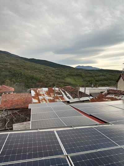 The image shows a view from a rooftop in what appears to be Petrovo, Bulgaria, judging by the architecture and mountainous terrain. The foreground is dominated by rows of solar panels, with some appearing cleaner and newer than others. Adjacent to the solar panels are older buildings with rusted metal roofs and tiled roofs of varying conditions. Beyond the buildings, a heavily forested mountain range stretches into the distance under an overcast sky. The overall scene suggests a juxtaposition of older and newer technologies in a rural or semi-rural setting.