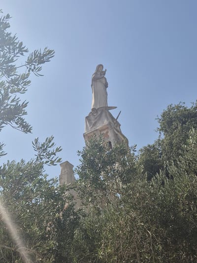 A stone statue of the Virgin Mary holding the infant Jesus stands atop a weathered stone spire, partially obscured by the branches of olive trees. The statue is visible against a clear blue sky. The spire appears to be part of a larger, ancient stone structure, with visible architectural details and signs of age. The foreground is dominated by the lush green foliage of olive trees, with sunlight glinting through the leaves. The image was taken outdoors, suggesting a daytime setting, likely in a location with abundant olive trees, such as Abu Gosh, Israel.