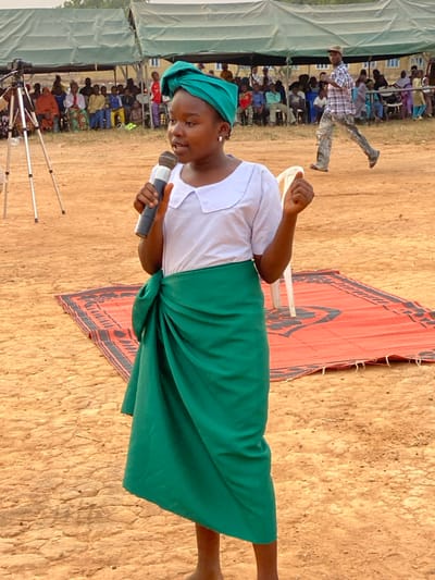 The image depicts an outdoor public event in Agunu, Nigeria, under bright daylight. A young person, likely a girl or young woman with dark skin, is the central subject, holding a microphone. She is dressed in a white short-sleeved top, a green headwrap, and a green fabric tied around her waist, serving as a skirt or wrapper. Her expression appears focused, suggesting she is speaking, singing, or delivering a presentation.
Behind her, a red and black patterned mat or rug is laid on the light brown, dusty ground. A white chair is partially visible behind the main subject. To the left, a large audience is seated in rows under light-colored tents or temporary shelters. One man in a plaid shirt and light pants is seen in motion in the middle background. Further back, some buildings are indistinctly visible. In the left foreground, several metal tripods or stands are set up, possibly for cameras or other equipment. The setting suggests a community gathering, performance, or public address. No visible text is present in the image.