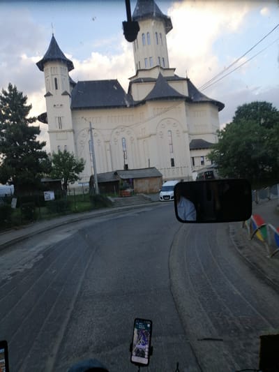 The image captures a view from a vehicle in Fălticeni, Romania, looking towards a large, cream-colored building with a dark roof, reminiscent of a church or monastery, featuring multiple spires and arched windows. The building is situated behind a road with a white van parked to its left. A side mirror of the vehicle is visible in the foreground, reflecting a person in a light-colored shirt. The road surface appears wet in places, with tire tracks visible. A smartphone displaying a navigation app is mounted on the dashboard. The sky is cloudy, with daylight fading. In the background, trees and smaller buildings line the street.