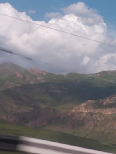 The image captures a panoramic view of a mountainous landscape under a bright, partly cloudy sky. The upper left portion of the frame is dominated by a clear blue sky interspersed with large, white, fluffy cumulus clouds.
The central and lower right sections of the image depict steep mountain slopes or large hills, densely covered with varied green vegetation, ranging from darker forest-like areas to lighter green patches. Exposed rock formations, displaying reddish-brown and earthy tones, are visible along the contours of the terrain, particularly on the left side of the prominent mountain feature.
A thin, light-colored cable or wire traverses the scene diagonally from the upper left to the lower right, crossing both the sky and the mountain. This, along with a blurry, light-colored vertical structure partially visible on the far right edge (likely a window frame or structural element), suggests the photograph was taken from an elevated perspective, possibly from a cable car or similar aerial transport.
The setting is an expansive outdoor natural environment, likely in a high-altitude or rugged region. The time of day appears to be daytime, indicated by the bright light and clear visibility. There are no people or discernible text visible. The weather is fair with scattered clouds. The scene reflects the natural beauty of mountainous terrain, consistent with the location context of Yelpin, Armenia.