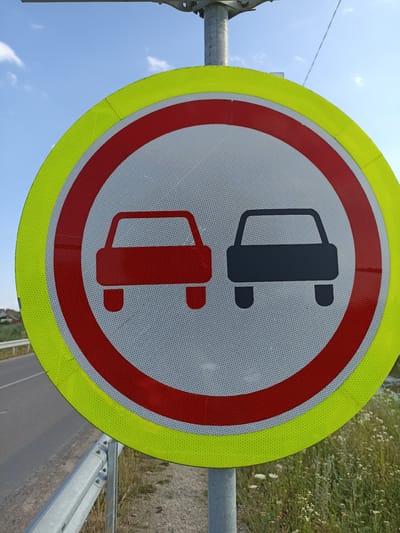 The image shows a close-up of a traffic sign in Zoziv, Ukraine. The sign is circular with a yellow outer border and a red inner border. Inside the red border, there are two car silhouettes. The car on the left is red, and the car on the right is black. The sign is mounted on a metal pole and is positioned next to a road with a metal guardrail. The background shows a blue sky with some clouds, a grassy area with wildflowers, and some distant trees. The sign appears to be a "No Overtaking" or "No Passing" sign for vehicles.