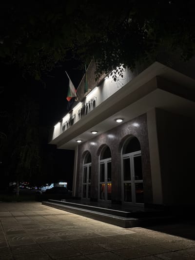 The image shows the exterior of a building at night, illuminated by external lights. The building appears to be a public institution, with flags displayed at the top. The architectural style features arched doorways and marble-like cladding. A paved walkway leads up to the entrance. In the background, a tree overhangs the building, and streetlights and a few vehicles are visible, suggesting an urban environment. The scene is set in the city of Straldzha, Bulgaria.