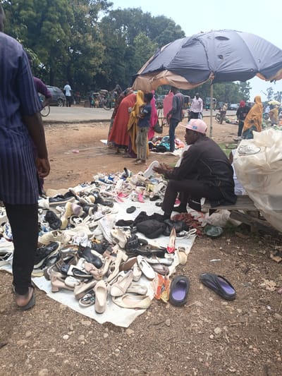 The image depicts an outdoor market scene, likely in Tilde, Nigeria, with a focus on footwear. In the foreground, a large pile of various shoes, including sneakers, sandals, and heels, is spread out on a white sheet on the ground. A man wearing a dark outfit and a pink cap sits on a wooden bench, examining a white sneaker. To the left, a person wearing a blue and purple striped garment and black pants stands with their back to the camera, appearing to observe the shoe display. Further back, other individuals are visible, some wearing traditional clothing, and a large, damaged umbrella provides shade over a section of the market. In the background, several tricycles, cars, and motorcycles suggest a busy thoroughfare. The ground is dusty and scattered with small stones and debris.