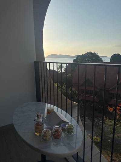 This image depicts a balcony scene overlooking the sea and distant islands in Cenang Beach, Malaysia, at either sunrise or sunset. In the foreground, an oval white marble-top table is positioned. On the table, from left to right, are a partially full bottle of "Monkey Shoulder" whiskey, a tall glass containing an amber liquid with ice, a folded white menu or pamphlet, and four small, clear glass jars with lids containing various snacks: light-colored items, reddish items, light brown items, and green items. The balcony features a plain light-colored wall on the left, a distinctive curved architectural wall segment, and a dark metal railing with vertical bars. Below the balcony, several buildings with distinct tiled roofs are visible, nestled among numerous green trees and foliage. A paved road or path is discernible below these structures. The background shows a calm body of water extending to multiple landmasses or islands on the horizon, beneath a pale blue sky that transitions to a warm yellowish hue, indicating the time of day. No individuals are present in the scene.