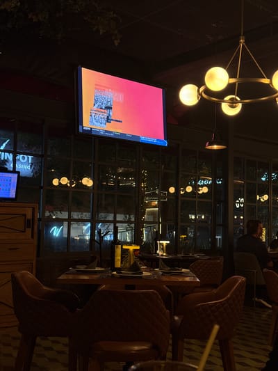 The image displays the interior of a dimly lit restaurant or bar at night, confirmed to be a WinPot Casino establishment in San Agustín, Mexico, indicated by a partially visible neon sign outside the window. A large flat-screen television, mounted high on a dark wall, is the central focus, displaying a sports graphic with a bright orange and pink gradient background. The ESPN logo is visible in the bottom left corner of the screen, along with text indicating an "AMISTOSO Brasil" versus "Francia" match, with a score of "BRA > Bremer (76')" and "1 Francia," concluding with "2 Final."

In the foreground, a dining table is set with dark placemats, white plates, dark folded napkins, and cutlery. A bottle of red wine and a small, rectangular golden table lamp illuminate the setting. The table is surrounded by four brown, diamond-quilted leather armchairs. Additional similar tables and chairs are visible in the background.

Overhead, a prominent brass-colored chandelier with six spherical, glowing white bulbs hangs, accompanied by a smaller, dark industrial-style pendant light. The ceiling is dark with a subtle grid pattern.

Large, dark-framed windows form an exterior wall, revealing reflections of interior lights and glimpses of the outdoor environment. Outside, neon signs are visible, including a white "W" and parts of the "WIN POT CASINO" marquee. Distant string lights and other urban lights contribute to the nighttime ambiance. On the far left, a wooden cabinet houses a smaller digital monitor displaying colorful graphics. A single individual is seated with their back to the camera at a table on the right side of the frame. A drinking straw is partially visible in the bottom right corner.