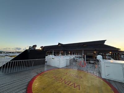 The image depicts a scene at dawn or dusk, viewed from the deck of a vessel, likely a ferry, docked at the port of Papeete, French Polynesia. In the immediate foreground, a grey metal deck with a non-slip textured pattern features a prominent yellow circular marking with a red border, displaying the inverted text "WINCH ONLY." A silver metal safety railing runs horizontally across the lower mid-frame. Two white, rectangular utility boxes are situated on the deck; the one on the left is marked "H11." A dark, upward-sloping structural element with two illuminated, pipe-like fixtures is visible on the left side of the deck.

In the midground, beyond the railing, stands a large building with a distinctive dark, multi-tiered, peaked roof, exhibiting traditional or tropical architectural characteristics. Several windows are visible, some of which are illuminated. A flagpole displaying what appears to be a French flag is positioned near the building. A red and white lifebuoy hangs from a railing in front of the building. The background consists of a clear sky, transitioning from a light blue overhead to a pale yellow-orange hue near the horizon. Distant port infrastructure and landforms are faintly discernible. No individuals or dynamic actions are depicted in the scene.