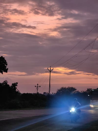 This is a vertical, eye-level, medium shot of a road scene in Dattapur, India, during twilight. The sky displays a gradient of pink and purple hues with scattered clouds. In the foreground, a dark asphalt road with a white line marking the edge is visible. A motorcycle, illuminated by its bright headlights, is approaching the viewer from the right side of the frame. The rider, wearing a helmet and a dark shirt, is silhouetted against the lights. To the left of the road, dense foliage and trees line the background, with two utility poles with electrical wires crisscrossing the sky. A large vehicle with its headlights on is partially visible to the right of the motorcycle, also heading towards the viewer. The overall atmosphere is dim, with artificial light from the vehicles creating bright, lens flare-like effects.