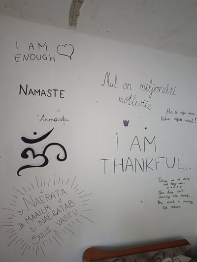 The image shows a wall with handwritten text and symbols. On the left side, in large block letters, it says "I AM ENOUGH" with a heart symbol next to it. Below that, in block letters, is "Namaste". Further down is the Om symbol, a religious icon in Hinduism. To the right of "Namaste" and above the Om symbol, in cursive, is "Armasta" with small hearts.
In the upper right section of the wall, there is text in cursive. The first line appears to be "Mul on miljonäri". The second line is "mõtteviis". Below this, in a smaller font, are the words "Ma ei vaja raha, Raha vajale mind!". There is also a small purple butterfly graphic.
Centered below the butterfly and to the right of the Om symbol, in large block letters, it reads "I AM THANKFUL...".
In the lower right corner, there is text in cursive: "Things are not always what they seem, **** The deer isn't crossing the road, the road is crossing. THE FOREST."
In the lower left section, there is text written in a sunburst pattern. The text reads: "Naerata" at the top, then "Ja maailm" below it, and "Naeratab sulle vastu" at the bottom. Small smiley face emojis are scattered among the text in this section.
The wall appears to be an interior wall, possibly in a home or community space. The ceiling above the wall shows signs of repair or unfinished work. A piece of furniture, possibly a chair or sofa, is visible at the bottom of the image. The presence of these handwritten messages and symbols suggests a personal or spiritual expression. There are no people visible in the image. The location in the city of Kohila, Estonia, is not discernible from the visual content of the image itself.