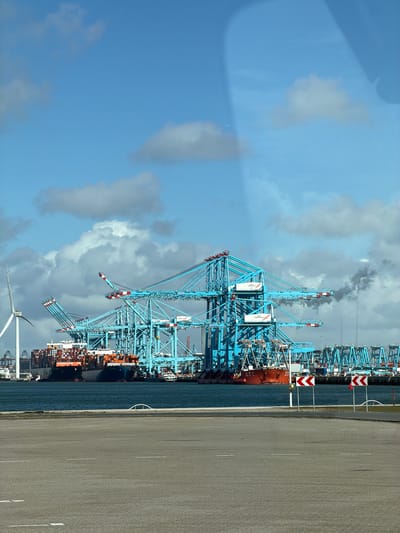 A wide-angle photograph depicts an industrial port scene at Maasvlakte Rotterdam, Netherlands, under a partly cloudy blue sky. The lower third of the image features an empty asphalt paved area with white dashed lane markings. Two red and white chevron signs, oriented to the left, stand on the right side of this paved area, preceding a body of dark blue-green water. Across the water, a large container terminal is active, dominated by numerous massive, bright blue gantry cranes, many marked with "APM TERMINALS" and featuring red and white details on their booms. Several large cargo vessels are docked: a container ship, heavily laden with stacked containers (predominantly red and orange), is visible on the left, and another vessel with a red hull is docked further right, with part of a name like "ZHUANG" visible. A plume of dark gray smoke rises into the sky from the industrial area to the right of the central cranes. On the far left, a tall white wind turbine is visible. No individuals are discernible. The scene is illuminated by natural daylight.