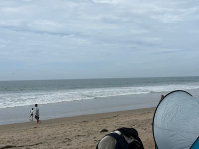 This image depicts a cloudy day at a beach in San Clemente, California, United States of America. The scene is characterized by a sandy beach with gentle waves lapping the shore. In the mid-ground, two individuals, an adult male and a child, are walking near the water's edge. The adult is wearing a light-colored t-shirt, shorts, and a baseball cap, and is carrying a pair of flip-flops. The child, also in light-colored clothing, is ahead of the adult, walking in the shallow water. In the far distance, a person can be seen in the ocean. In the foreground, a partially visible beach tent or shelter is present, with some clothing items placed on the sand nearby. The sky is overcast with scattered clouds, and the ocean appears a muted green-blue.