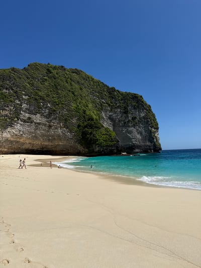 The image depicts a tropical beach scene in Bunga Mekar, Indonesia. A large, steep, rocky cliff covered with green vegetation rises sharply from the sand. To the right of the cliff, a sandy beach stretches along the turquoise ocean. Several people are walking along the shoreline, and one person appears to be swimming in the water. Footprints are visible in the sand. The sky is a clear, bright blue.