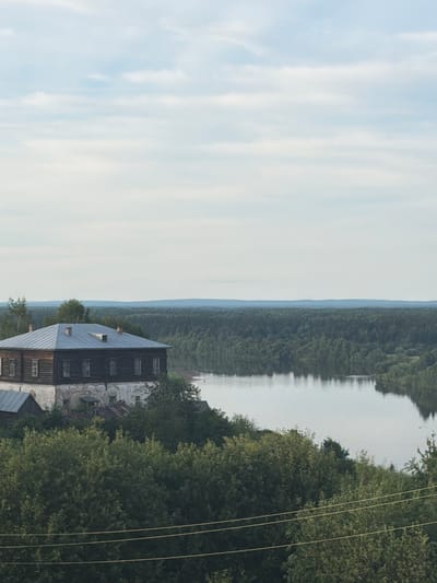 The image depicts a pastoral scene with a prominent wooden building, likely a traditional Russian house or barn, situated on the edge of a tranquil river. The building features a dark, weathered wood exterior and a sloping metal roof with visible windows. Adjacent to the building, lush green trees form a dense backdrop, extending towards the riverbank. The river itself appears calm, reflecting the overcast sky and the surrounding foliage. In the distance, a hazy line of hills or mountains can be seen, shrouded in a light mist, suggesting a cool or humid atmosphere. The sky is a soft, pale blue with streaks of white clouds, indicating it's likely daytime, possibly late afternoon or early morning due to the diffused light. The overall mood is serene and somewhat rustic, evoking a sense of quiet rural life. Power lines are visible in the foreground, crossing diagonally across the frame, hinting at the presence of modern infrastructure in this otherwise traditional setting. The image appears to be taken from a slightly elevated position, offering a panoramic view of the landscape. No people are visible in the scene, and there is no visible text.