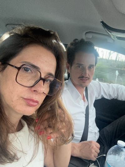 A selfie-style photograph shows two individuals inside a vehicle. The woman in the foreground wears glasses and a light-colored garment. Behind her, a man in a white shirt is partially visible, wearing a seatbelt. A bottle of water is present near the man. The background appears to show an exterior scene possibly in Alcobaça, Portugal. The car interior is visible with a headrest and handle. The image is slightly angled.