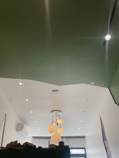 The image shows an interior view of a modern space with a high ceiling. The ceiling is painted in two contrasting colors: a muted green on the upper section and white on the lower section. The white section of the ceiling is angled and appears to have a wavy, organic edge where it meets the green. Several recessed ceiling lights are visible in the white section. A contemporary chandelier with multiple spherical glass globes hangs from the white ceiling. An air conditioning unit is mounted on a white wall. In the foreground, on a dark surface, there are several brown paper bags, some of which are crumpled. The environment suggests a commercial or public space, possibly a cafe or a shop. The provided information states the location is in Meaux, France, but this is not visually discernible from the image itself. There are no people or discernible actions visible in the image.