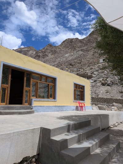 The image shows a yellow building with wooden framed windows and doors, situated in a mountainous, arid environment. A red plastic chair is placed on the concrete patio in front of one of the windows. Steps lead down from the patio to a sandy ground. The background features a rocky, barren mountain slope with scattered boulders and sparse vegetation. Prayer flags are visible on the hillside, suggesting a spiritual or cultural significance to the location. The sky is bright blue with scattered white clouds. The scene appears to be in Panamik, India, a region known for its natural hot springs and Buddhist monasteries.