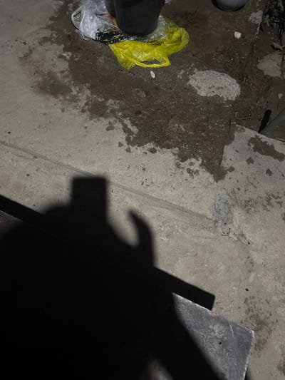 The image depicts a close-up, low-angle shot of a damp concrete surface with scattered debris. In the upper portion of the frame, several plastic bags, one yellow and one white, are visible. The white bag appears to contain dark objects and is partially transparent. A dark, cylindrical object, possibly a bucket, is positioned next to the bags. The concrete is wet and discolored in patches, suggesting recent exposure to liquid. A distinct shadow is cast across the lower left portion of the image, forming an abstract shape that resembles a hand and a rectangular object. This shadow is a prominent feature, obscuring a portion of the concrete. The overall environment appears to be an outdoor, possibly urban, setting, given the concrete surface and discarded plastic. No specific landmarks or indicators of the city of Kojomkul, Kyrgyzstan are present in the image. The lighting suggests it may be nighttime or a dimly lit area, as evidenced by the strong shadows.