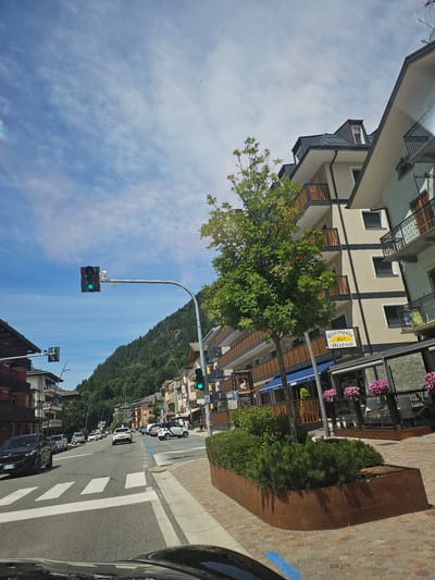 The image depicts a street scene in Aprica, Italy. The foreground shows the dashboard and windshield of a car, suggesting the photo was taken from a moving vehicle. The street is paved and has white pedestrian crossing lines. Traffic lights, with green lights illuminated, are visible. On the right side of the street are multi-story buildings with balconies, some adorned with flowers. One building has a sign that reads "Ristorante Bar Bistrot". A lush green tree stands in front of these buildings, and further down the street, a mountain covered in dense green forest rises under a blue sky with scattered white clouds. Several cars are on the road, including a black car on the left and a white car on the right. The overall impression is a sunny day in a town nestled beside a mountain.