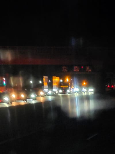 This is a blurry, low-light photograph taken at night on a road in Obertshausen, Germany. The image depicts a line of semi-trailer trucks, their headlights and taillights creating streaks of light due to motion blur. The trucks are on a paved road, and there are reflections on the wet surface, indicating it may have recently rained. Above the trucks, a reddish-brown structure with illuminated lettering is visible, likely a bridge or overpass. The overall scene suggests a highway or industrial area at night. There are no clearly discernible individuals or specific actions taking place, other than the vehicles in transit.
