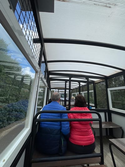 The interior of a multi-car, open-sided transport vehicle, located in Sant Llorenç des Cardassar, Spain, is depicted from a rearward perspective looking forward. Two individuals occupy the immediate foreground, seated and observed from behind. On the left is a male with short grey hair, wearing a blue and dark blue puffer jacket. On the right is a female with dark brown hair, wearing a red puffer jacket. Further down the vehicle, a third, lighter-haired individual is partially visible. The vehicle features a translucent, corrugated white roof and a dark metal framework with white lower panels. The left side is equipped with large, clear windows reflecting green trees and bushes under a blue sky with white clouds. The right side includes sections with dark wire mesh and smaller rectangular windows, revealing dense green foliage outside. Seating consists of dark bench-style seats, separated by black metal railings. Small, dark tables are affixed to the right side of the vehicle.