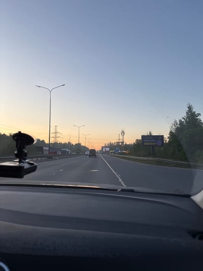The image captures a view from inside a moving vehicle on a multi-lane highway, looking forward and slightly right. The perspective is from the driver's or front passenger's seat, with part of the dashboard and windshield visible on the right and bottom edges, along with what appears to be a dashcam mount in the lower center.
The sky exhibits a clear, pre-dawn or post-dusk glow, transitioning from light blue at the top to a warm orange-yellow closer to the horizon, indicating either sunrise or sunset. Trees and dense vegetation line both sides of the highway, separating it from the surrounding landscape. Numerous street lights with dual arms extend along the roadside, and several large power lines with their supporting towers are visible in the distance, running parallel to the road.
Vehicles can be seen on the highway, both ahead of the observer and in adjacent lanes, suggesting moderate traffic. One prominent billboard is visible on the right side of the road, displaying text in Russian: "ЗАБОРЫ И НАВЕСЫ" (ZABORY I NAVESY), which translates to "FENCES AND CANOPIES." Below this, a phone number, "8-900-650-70-10," is partially visible. The scene depicts a typical drive along a main road or highway in the early morning or late evening, consistent with an area like Zanevka, Russia, given the Cyrillic text.
