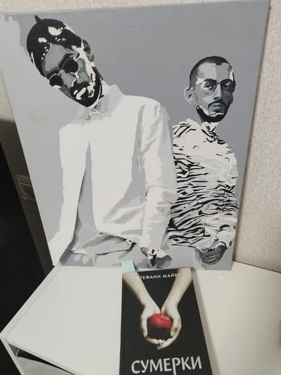 This is a photograph of a painting and a book displayed on a white table. The painting, rendered in grayscale, depicts two figures. The figure on the left is closer to the viewer and leans forward, his face obscured by dark, abstract shapes. He is wearing a white shirt. The figure on the right is further back, wearing glasses and a patterned shirt with dark stripes. The book is black with a red apple on the cover, and text in Russian. The painting is propped up against a light gray wall. The overall setting suggests an indoor display, possibly a home or an art exhibition. The image does not contain enough information to place the scene in Novostroika, Russia, or any specific city.