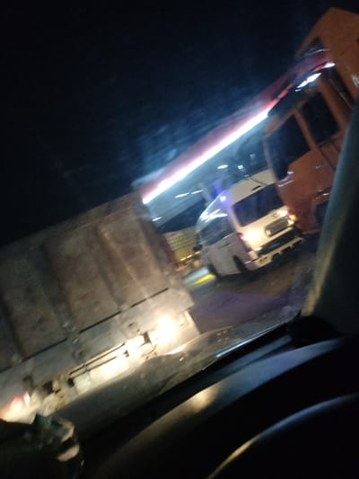The image depicts a nighttime scene with multiple vehicles, likely in a traffic or service area, possibly at a gas station due to the overhead lighting. A large, light-colored truck with a cargo container is on the left. To the right of it, a white van is visible, possibly a commercial vehicle, with its brake lights illuminated. Further to the right, an orange-colored truck or bus is parked. The environment appears to be a well-lit area despite the darkness, suggesting it's a public space with artificial lighting. The image quality is low, with motion blur and low light conditions. There is no indication of the specific location within Keudebirem, Indonesia.