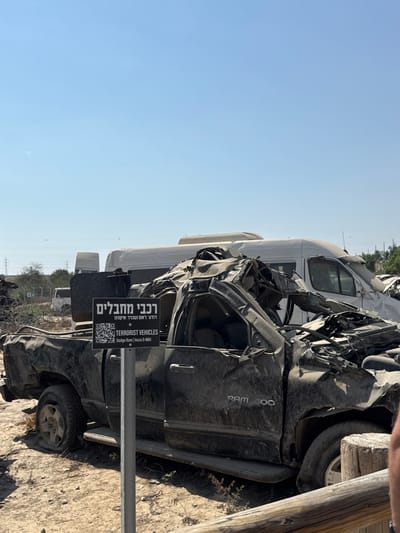The image displays an outdoor scene under a clear blue sky, located in the Sdot Negev Regional Council, Israel. The foreground features a severely damaged, dark-colored pickup truck, identified by its visible 'RAM 300' emblem as a Dodge Ram. The vehicle's front end is heavily mangled and collapsed, with the roof significantly caved in and ripped apart. The driver's side door appears open or missing its upper frame. Adjacent to the truck, a black rectangular sign on a grey metal post displays text in Hebrew and English, reading "רכבי מחבלים" (Vehicles of terrorists) and "TERRORIST VEHICLES," along with a QR code and further specifications "Dodge Ram | Isuzu D-MAX." Behind the pickup truck, a large white van, also extensively damaged with a crushed front section, is visible. Further in the background, a barbed wire fence separates additional vehicle debris and other indistinct structures. The ground is dry, dusty earth with sparse vegetation. Power lines are visible in the distant background. On the far right, a partial view of an individual's lower arm and shoulder is visible, positioned behind a wooden barrier.