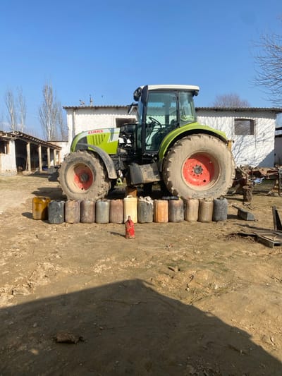 This image depicts a lime green and white CLAAS agricultural tractor parked on an unpaved dirt surface in Gulbahor, Uzbekistan. Approximately fourteen plastic jerrycans of various colors (including yellow, black, brown, gray, and orange) are arranged in a horizontal line on the ground directly in front of the tractor's left front and rear tires. A small, red, cylindrical object rests on the ground in front of the containers. In the background, an open-sided structure with wooden posts and a sloped roof is visible to the left, alongside a white, single-story building with small openings, upon whose roof several birds are perched. Bare deciduous trees are present in the mid-ground. The sky is clear and blue, indicating daytime. No people are visible.
