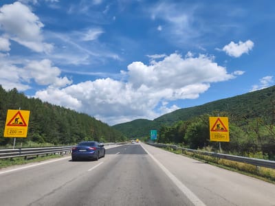 The image shows a view from inside a vehicle traveling on a highway. The road is paved and has two lanes in each direction, separated by a median. On the left side of the road, there is a blue car driving ahead. There are guardrails along the side of the road and a line of trees on the left. On the right side of the road, there is a hill covered in trees. The sky is blue with white clouds.
There are two yellow diamond-shaped warning signs with black pictograms and text. Both signs depict a worker with a shovel and a pile of dirt, indicating road work ahead. Below the pictogram on both signs, it reads "2000 m", suggesting that road work is approximately 2000 meters away. One sign is located on the left side of the road and the other on the right side.
A green highway sign is visible in the distance on the right side of the road, indicating directions to various locations, including "Potop" and "Express Road". This suggests the location is on a highway leading to or passing through Potop, Bulgaria.