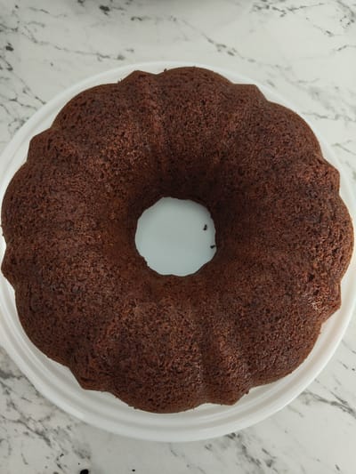 A dark chocolate bundt cake rests on a white cake stand, which is placed on a marble countertop. The cake has a rich, dark brown color and a textured surface. The bundt pan's fluted design is visible around the outer edge of the cake. The marble countertop has a white base with grey veining. No people are present in the image.